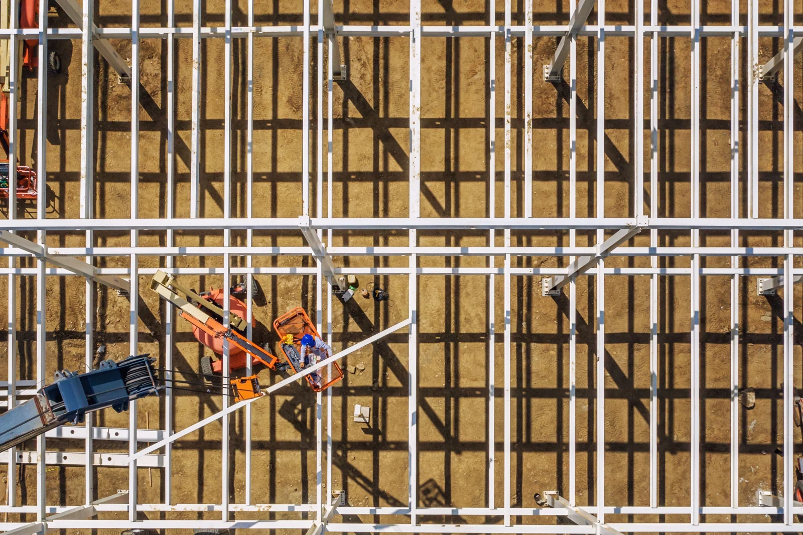 Steel roof framework viewed from above, with an orange boom lift below.