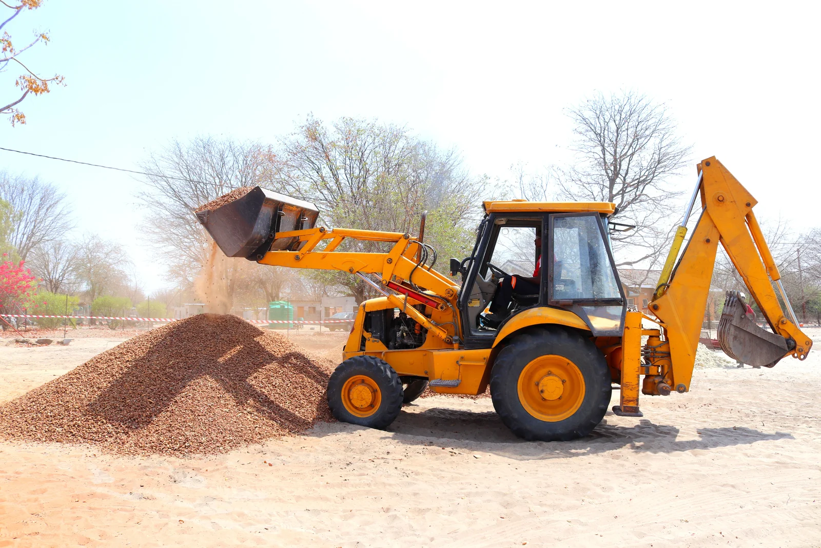 Yellow backhoe loader tipping aggregate onto a pile in daylight.