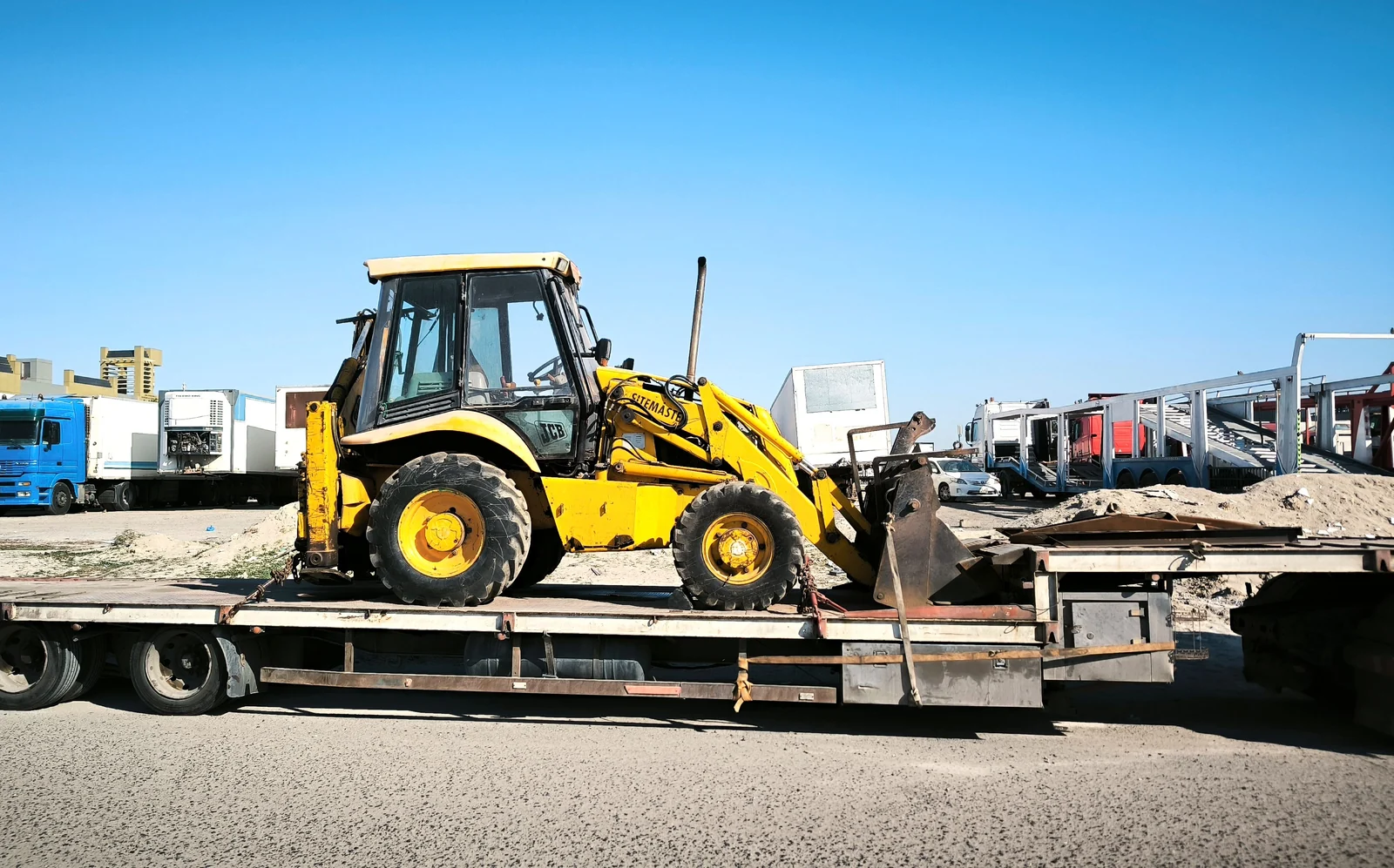 Yellow backhoe loader secured on a flatbed trailer in a transport yard.