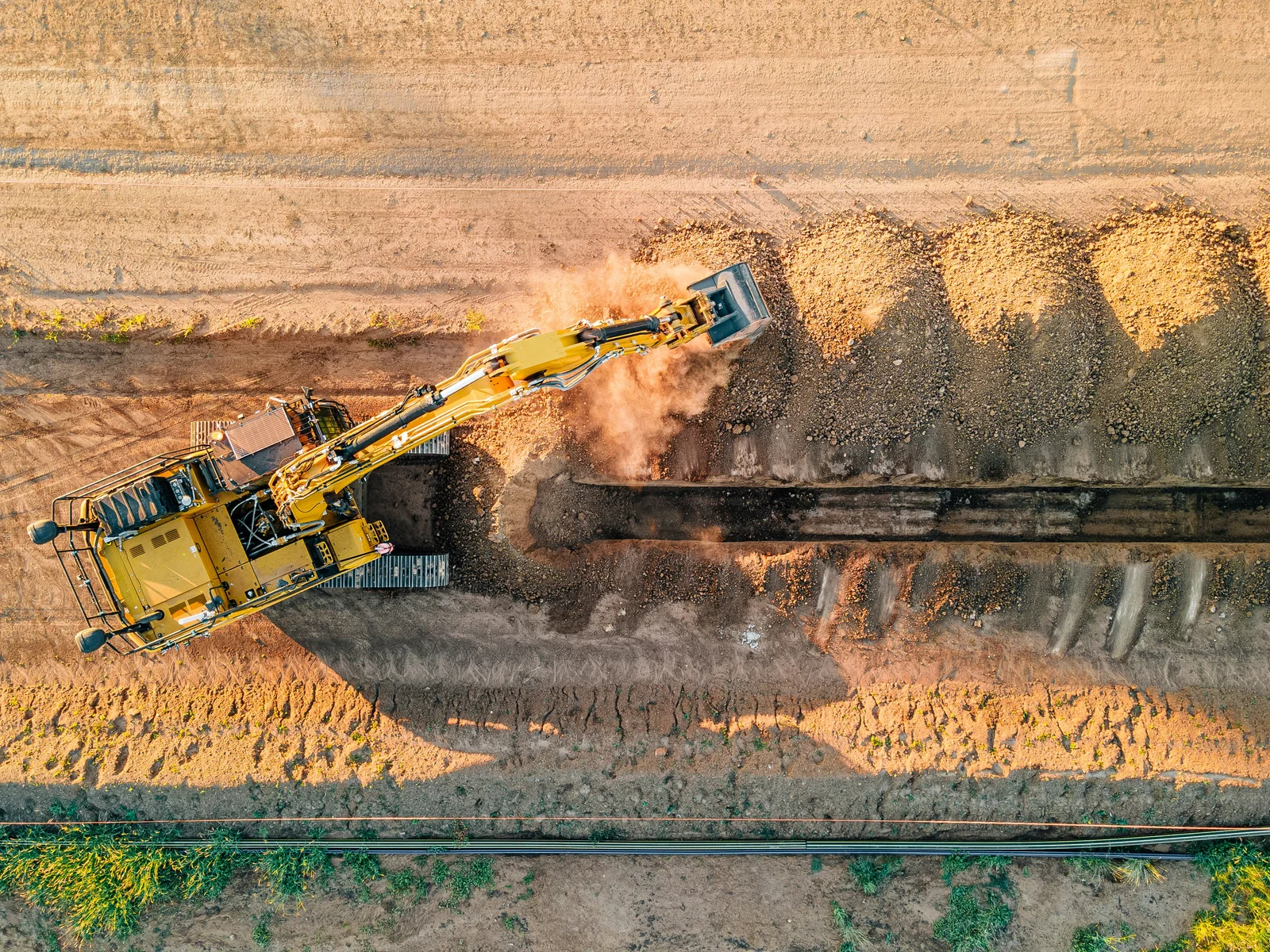 Overhead view of an excavator digging a straight trench in open ground.